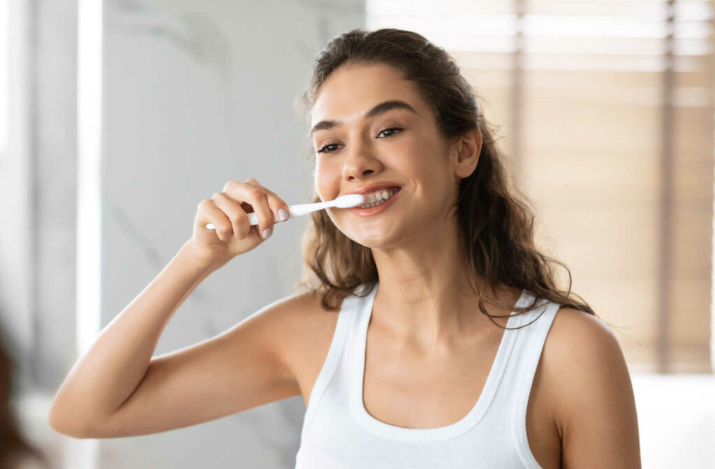 A woman holding a toothbrush in her hand and brushing her teeth in a white tank top.